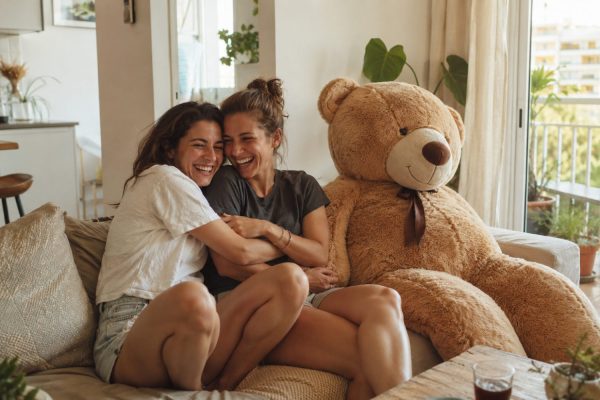 שתי חברות ילדות מחובקות וצוחקות בסלון ליד דובי ענק חום בגובה מטר ושמונים